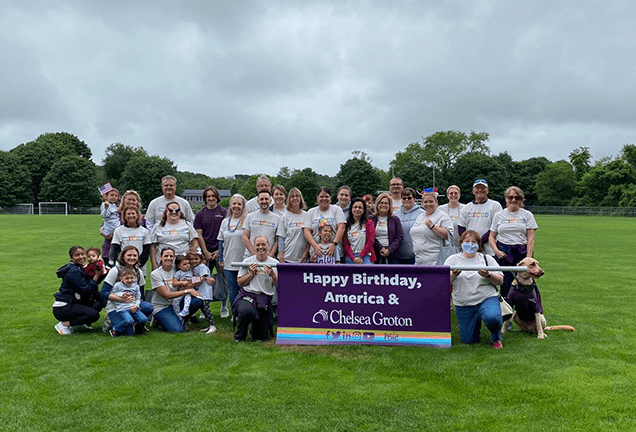 30 smiling people of varied ages wearing white tshirts on a soccer field  hold a large purple banner which heads "Happy Birthday, America & Chelsea Groton" in white text