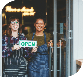 Two smiling women standing in a door frane hold a green side which reads "OPEN"