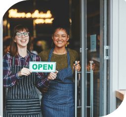 Two smiling woman who have leveraged small business banking stand in a doorway and holding a green open sign 