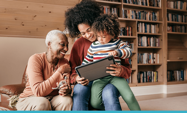 A smiling grandmother and mother with a toddler on her lap look at a tablet inside a library