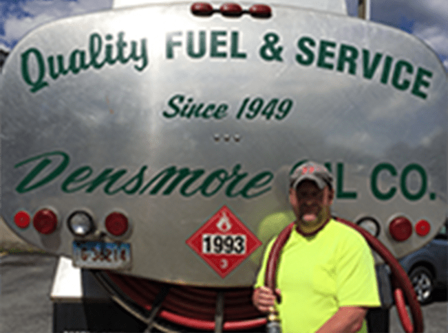 A man with a neon yellow hat stands in front of a steel fuel tank that reads "Quality Fuel & Service Since 1949" and holds a red hose on his shoulder