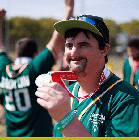 A smiling special olympics participant in green showing off a gold metal with a red ribbon around his neck