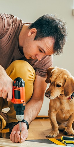 A man in a brown shirt uses an electric drill on the ground while a puppy watches