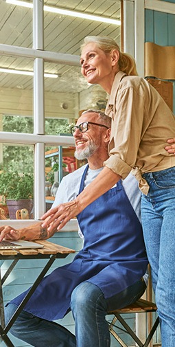 A smiling sitting older man wearing a blue apron with his arm around a smiling standing older woman looking at a laptop together