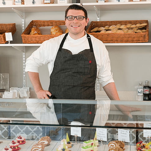 A male with glasses wearing in a white shirt and dark grey apron smiling behind a pastry counter