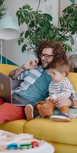A father sitting on a yellow couch and looking at a laptop has his arm around a toddler and talks on the phone