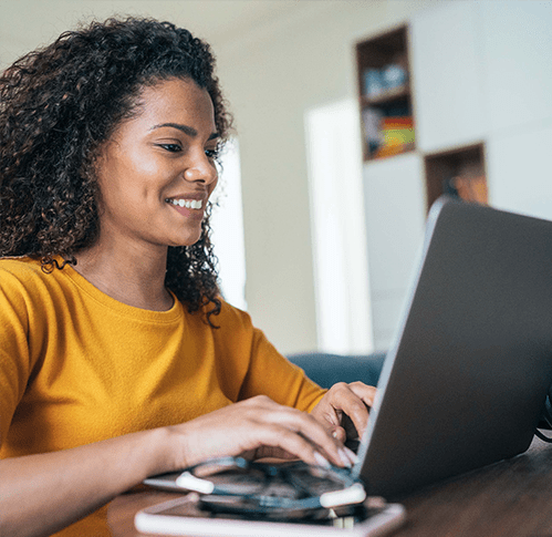 A smiling black woman wearing a yellow shirt uses a laptop